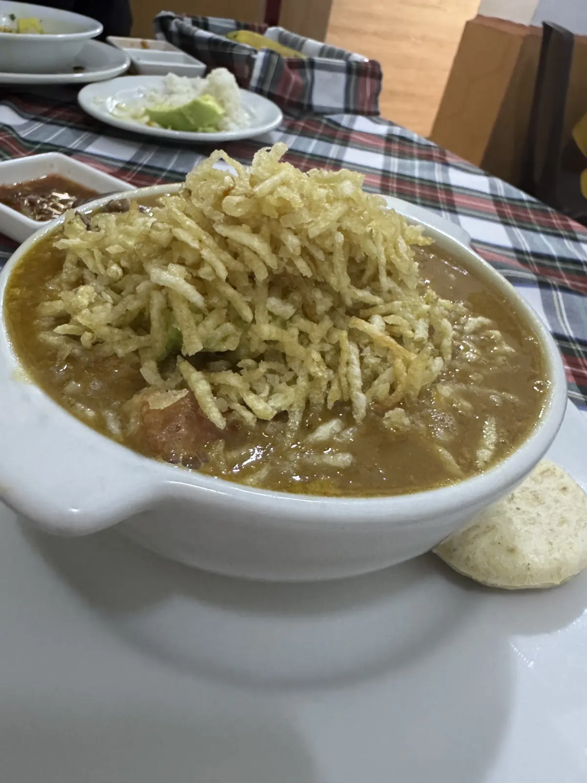 A bowl of soup topped with crispy fried potato straws, with a small arepa on the side, on a checked tablecloth at Ajiacos y Mondongos