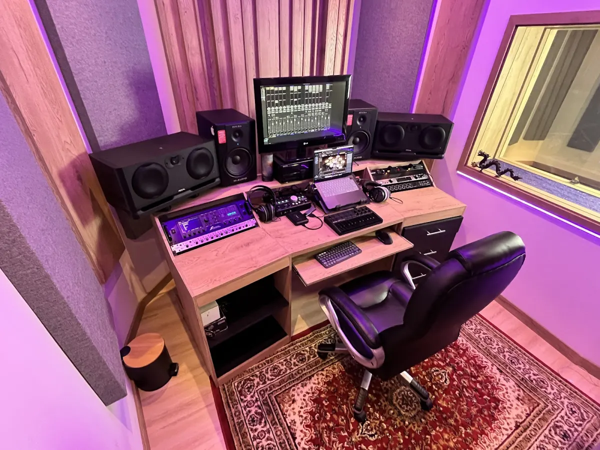 The control room at Arketipo Estudio, a wooden desk with studio monitors, a screen showing a mixing session, an audio interface and headphones, lit in purple beside the window into the recording room