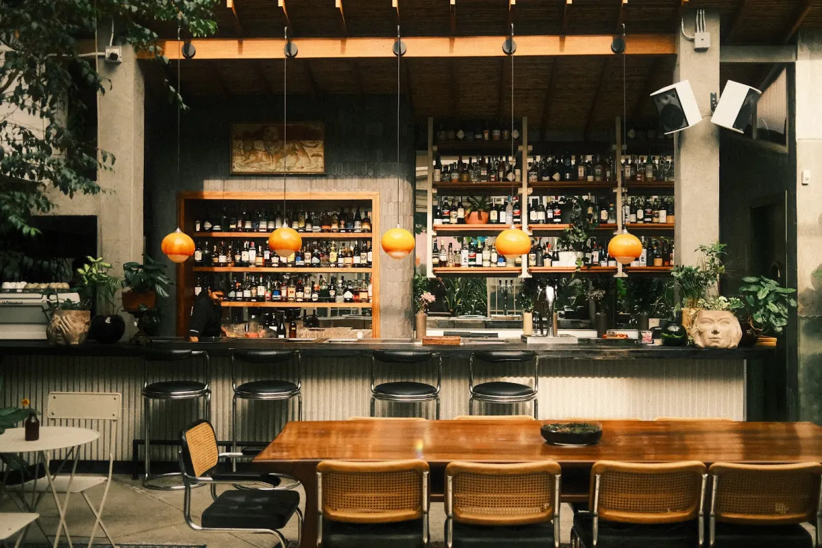 The bar at Café Dragón, with shelves of bottles, orange glass pendant lamps, black bar stools and a long wooden table with cane chairs