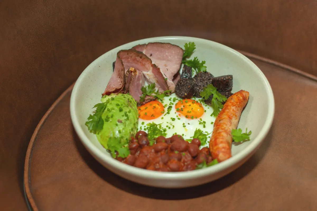 A bowl of two fried eggs, sliced ham, black pudding, a sausage, beans and avocado at Café Dragón