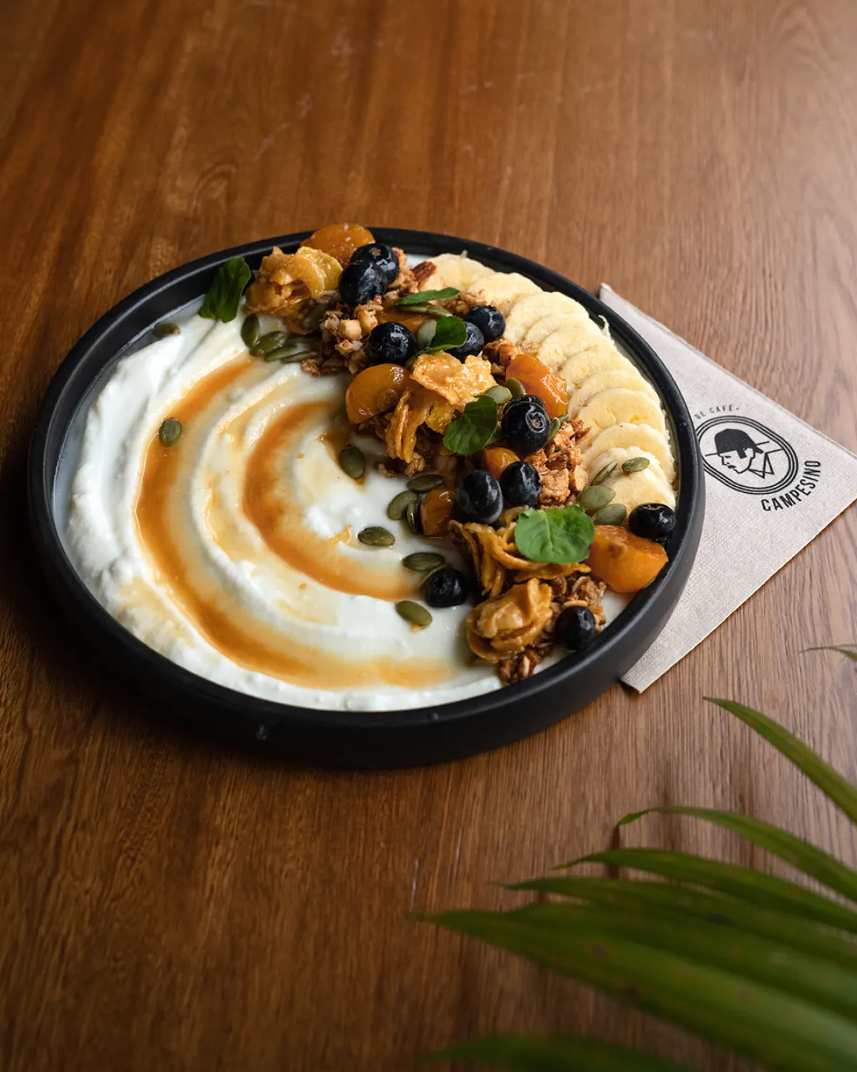 A plate of yoghurt with caramel sauce, granola, blueberries, banana slices and pumpkin seeds beside a Campesino Café napkin