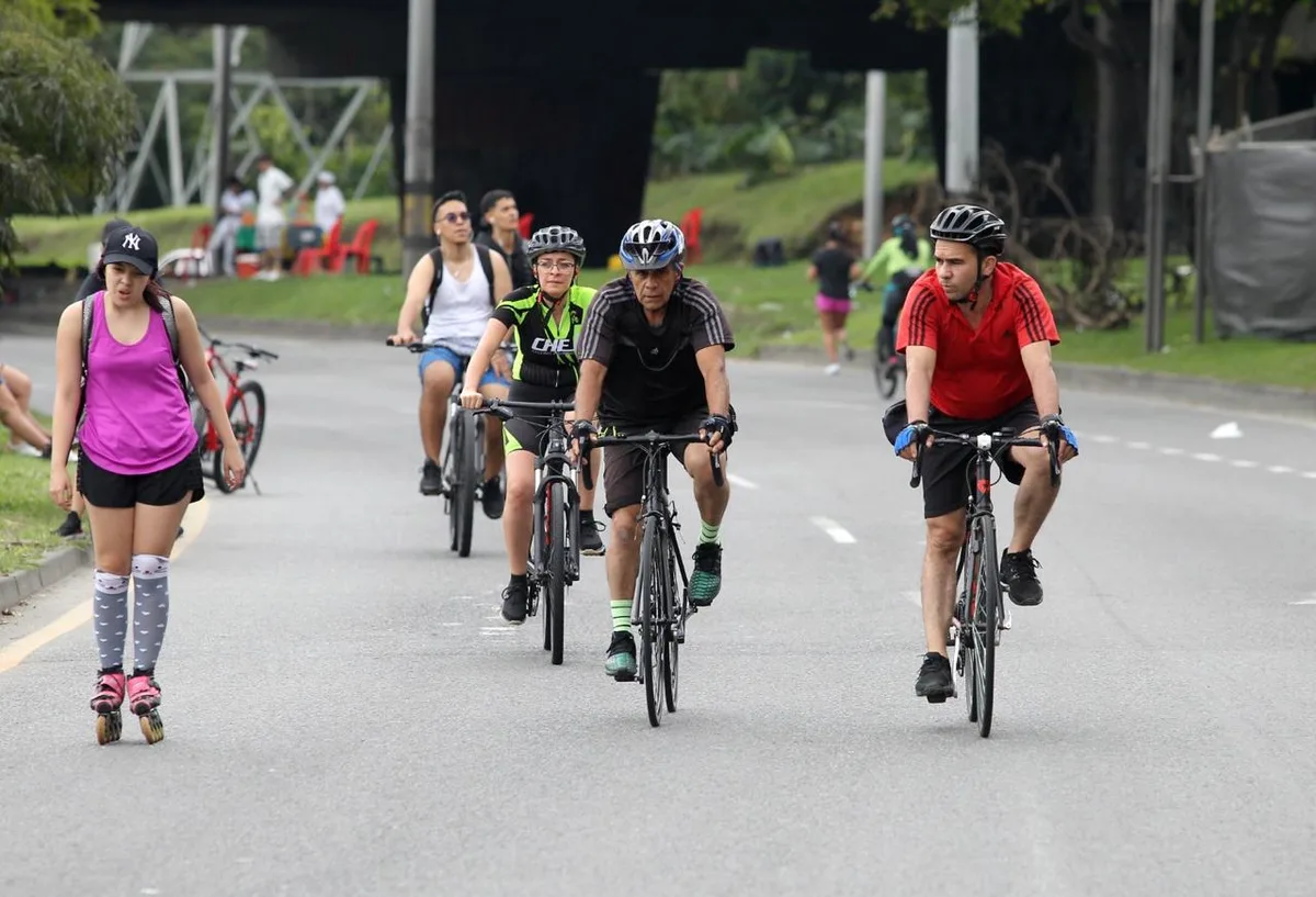 Cyclists and a roller skater sharing a wide car-free road on the Medellín Ciclovía, with trees and a footbridge behind