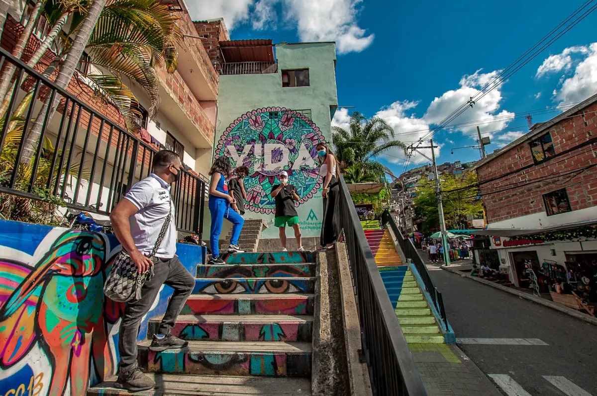 Visitors on a staircase painted with eyes and colourful patterns in Comuna 13, beside a green house with a large mandala mural spelling VIDA and a rainbow-painted stair beyond