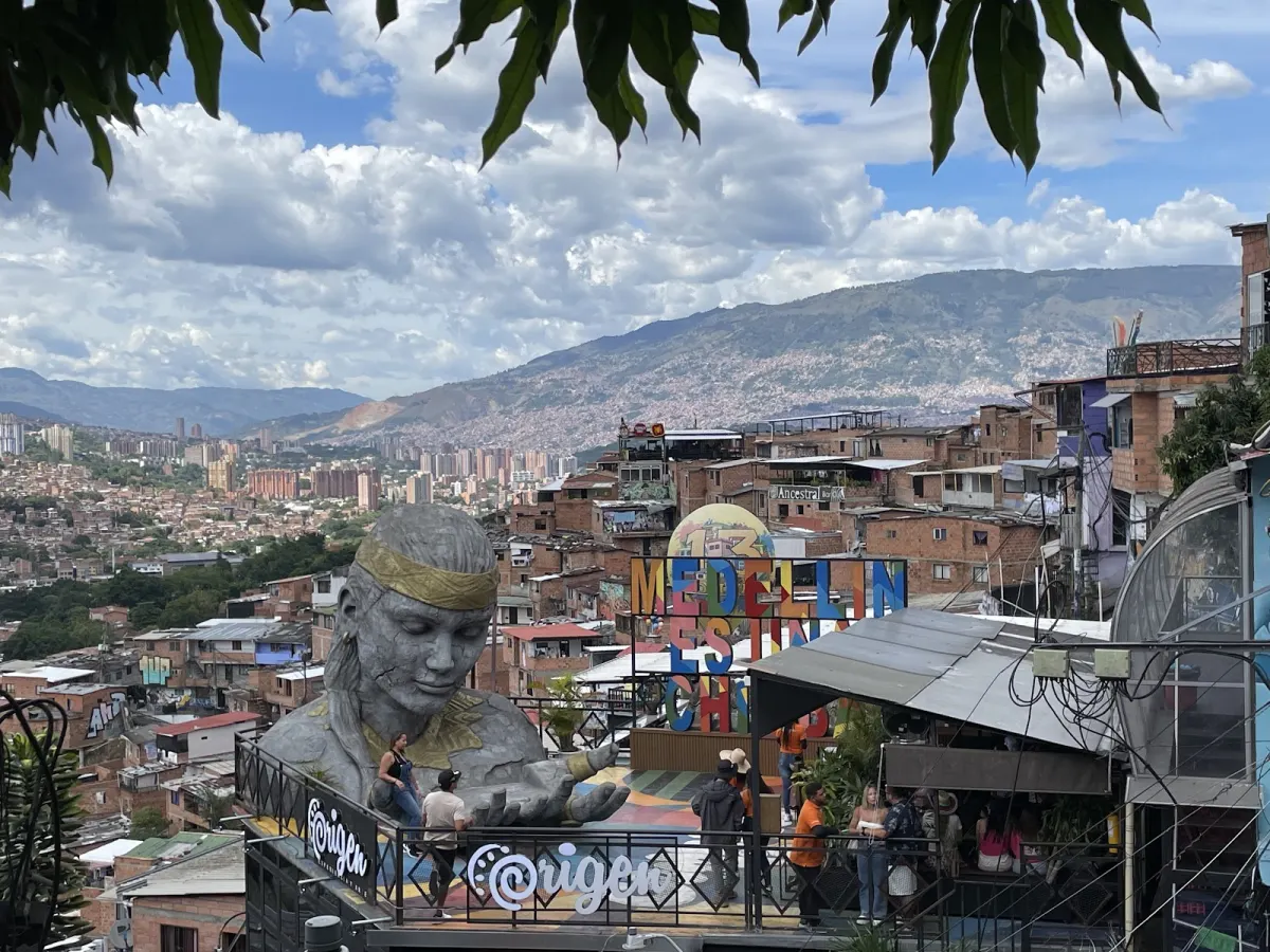A large grey sculpture of a woman's head and hands on a terrace in Comuna 13, with a colourful Medellín sign and the brick hillside neighbourhoods and city beyond