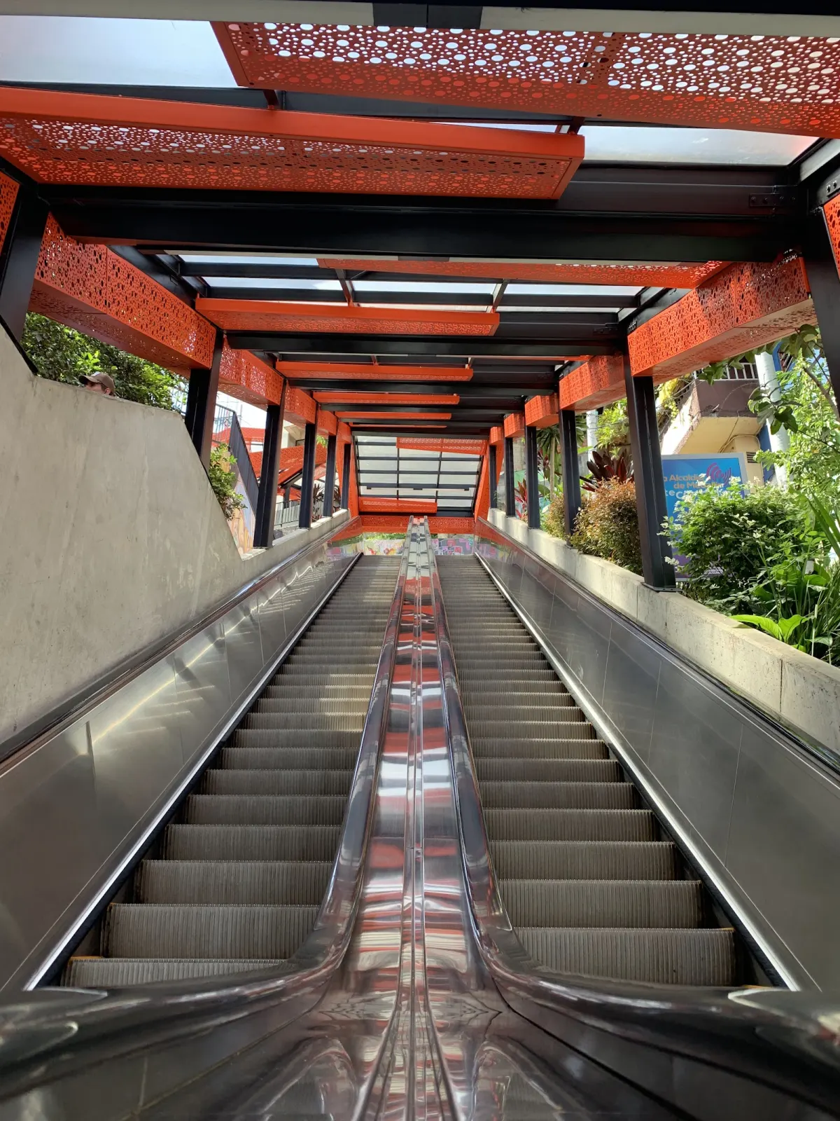 Looking up a pair of silver escalators under an orange perforated-metal roof, with plants along both sides