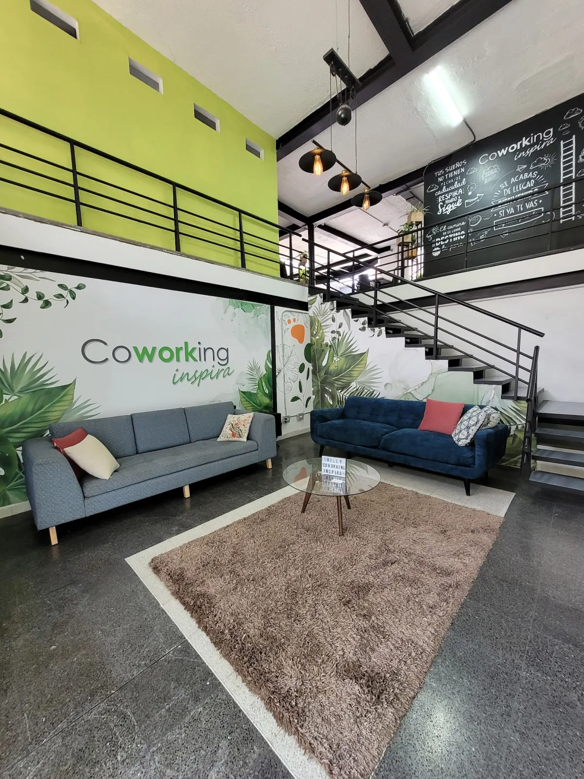 The double-height lounge at CoWorking Inspira, with two sofas on a rug in front of a leafy Coworking Inspira mural and a steel staircase up to a mezzanine