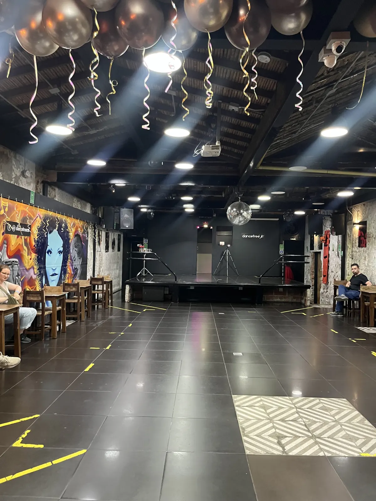 DANCEFREE's main room in El Poblado: a wide tiled dance floor under spotlights and gold balloons, a small stage at the far end, a mirror ball and a painted mural along the left wall.