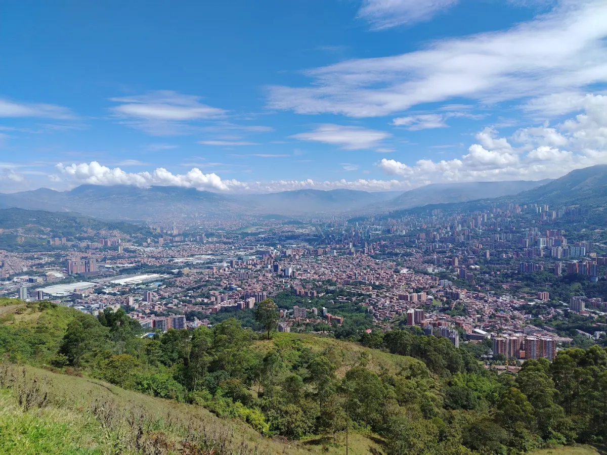 Daytime view from the green hillside at El Asfixiadero over the Aburrá Valley, the city spread across the valley floor below mountains and white clouds