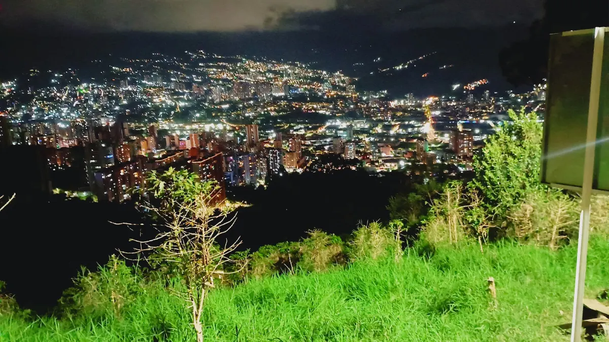 Night view from a grassy slope at El Asfixiadero over the lit-up city in the valley, with a signboard at the right edge