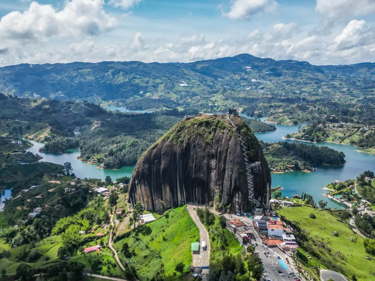 Aerial view of El Peñón de Guatapé rising beside the reservoir, its staircase up one side, the road and car park at its base and green hills and islands behind