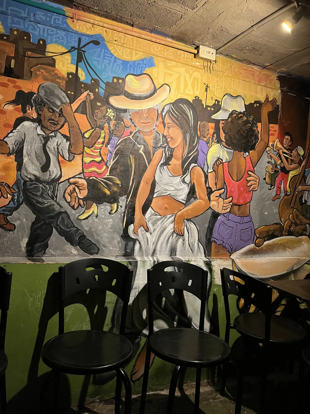 A painted mural of dancers and musicians in a street scene along a wall at El Tíbiri, above a row of black bar stools.