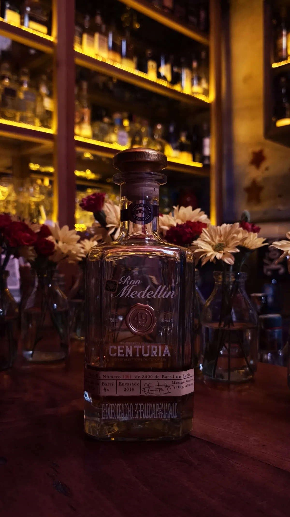 A bottle of Ron Medellín Centuria on the bar at ERRE, with flowers in vases and lit shelves of bottles behind