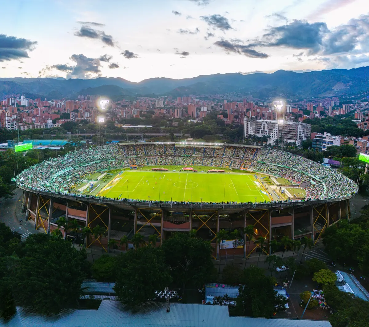 Aerial view of the Estadio Atanasio Girardot at dusk, its oval stands full of green-clad fans around a floodlit pitch, with Medellín's buildings and mountains behind