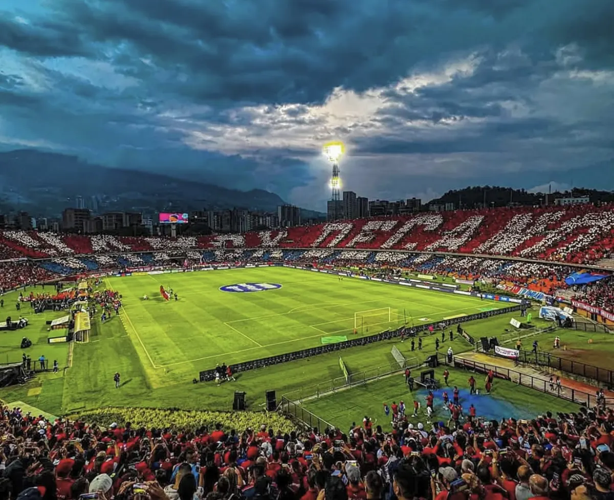 A crowd in red at the Estadio Atanasio Girardot under a stormy sky, the stands opposite forming a red, white and blue mosaic as players walk onto the pitch