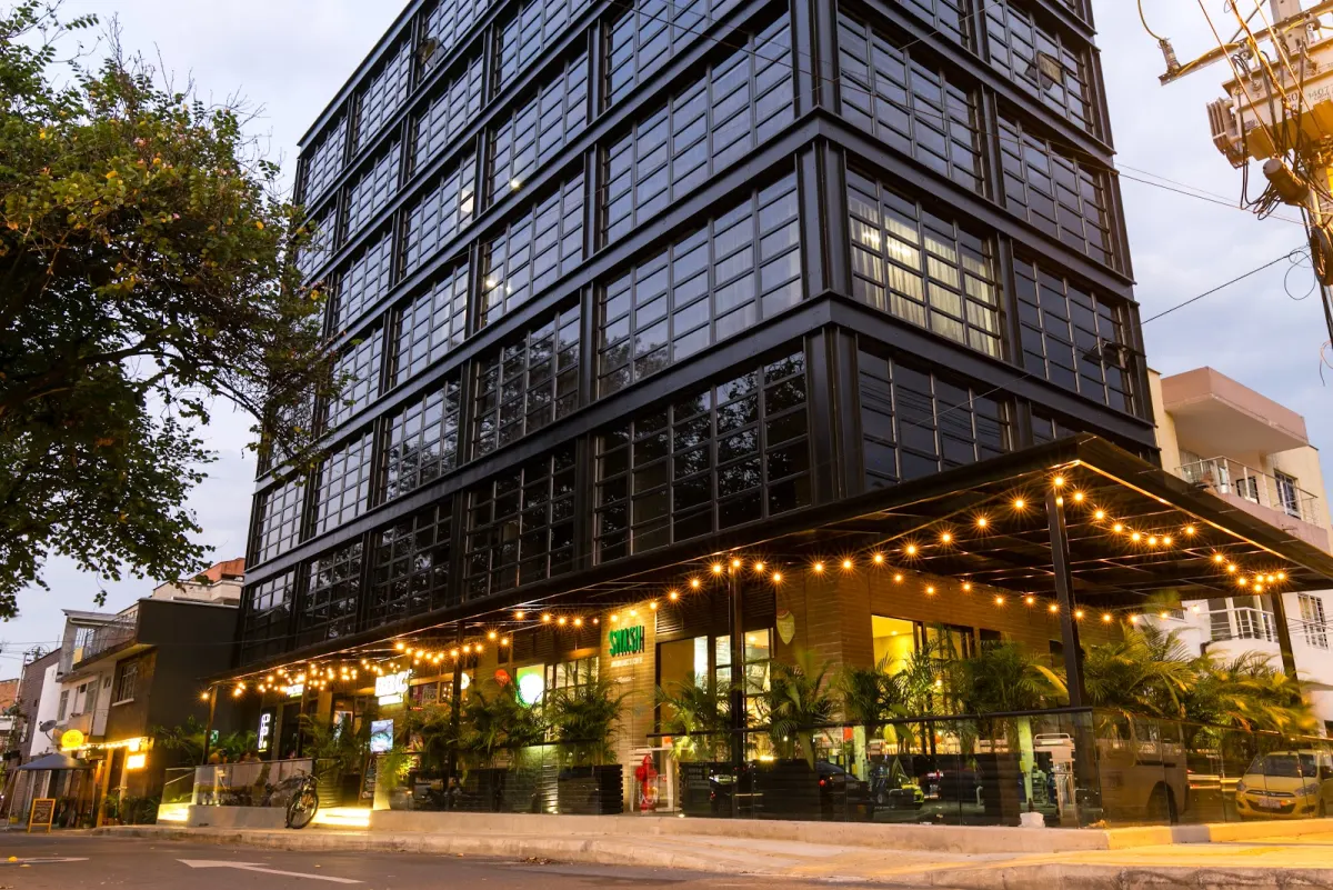 The black steel-and-glass Factory Lofts + Coworking building in Laureles at dusk, with string lights over the ground-floor terrace and the Smash restaurant sign