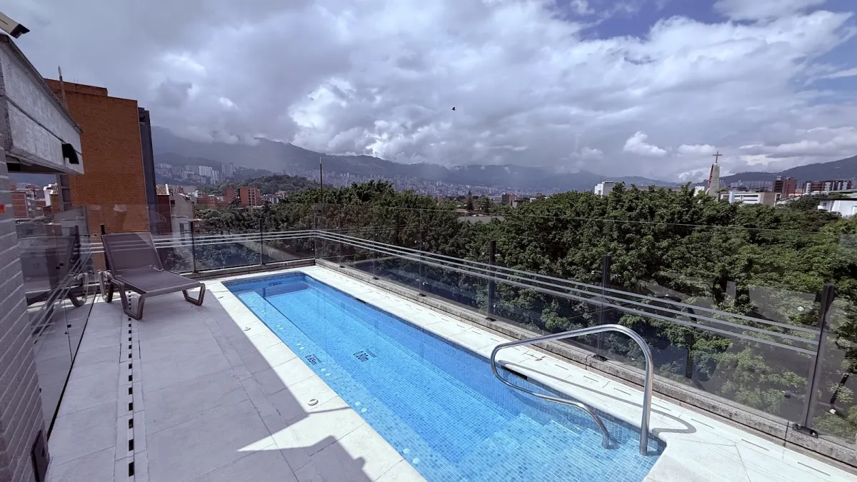 The narrow rooftop pool of the Factory Lofts + Coworking building, with a sun lounger, a glass railing, treetops and a view across Medellín to the mountains