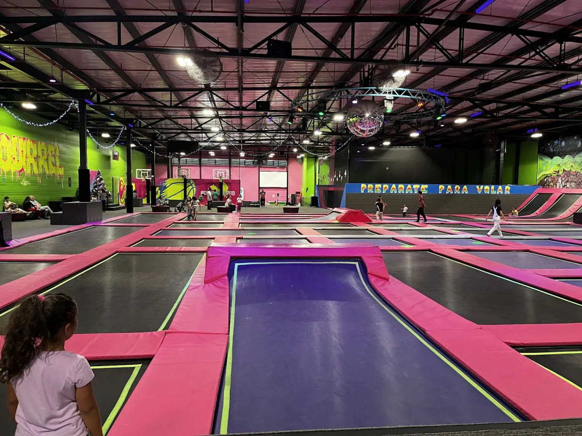 A long run of pink and purple trampolines under a high industrial roof, with a sign reading PREPARATE PARA VOLAR on the far wall