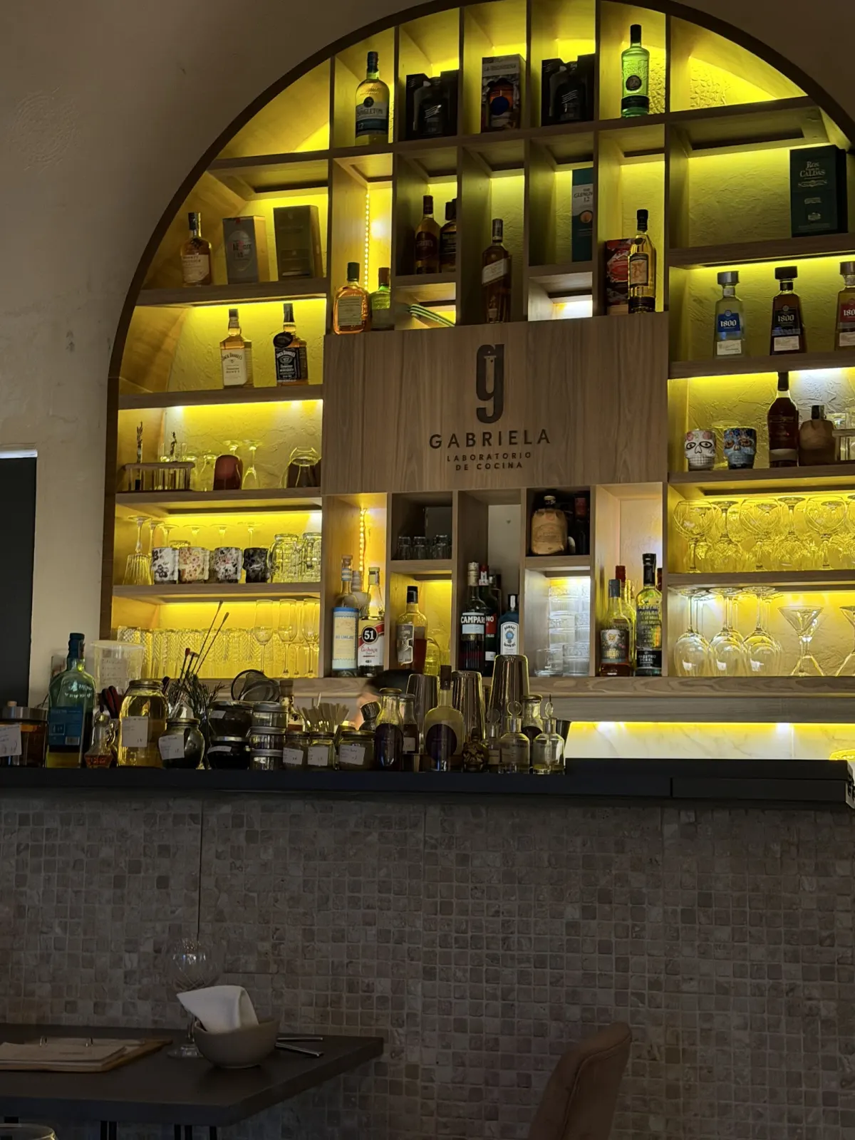 The bar at Gabriela Laboratorio de Cocina: an arched, yellow-lit back bar of bottle shelves around a wooden panel carrying the Gabriela logo