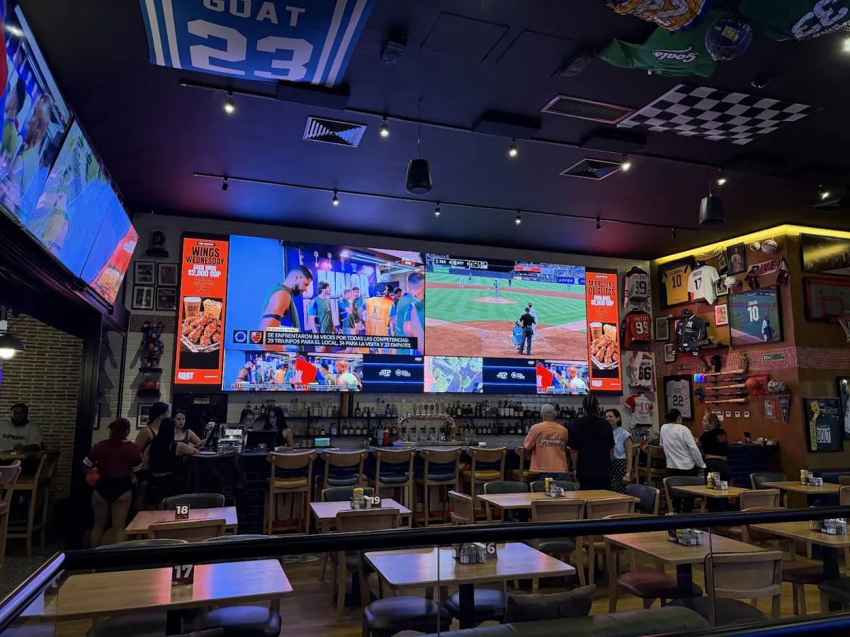 Main room at GOAT Sports Bar: a wall of large screens showing football and baseball above the bar, framed jerseys on the right wall and wooden tables in front