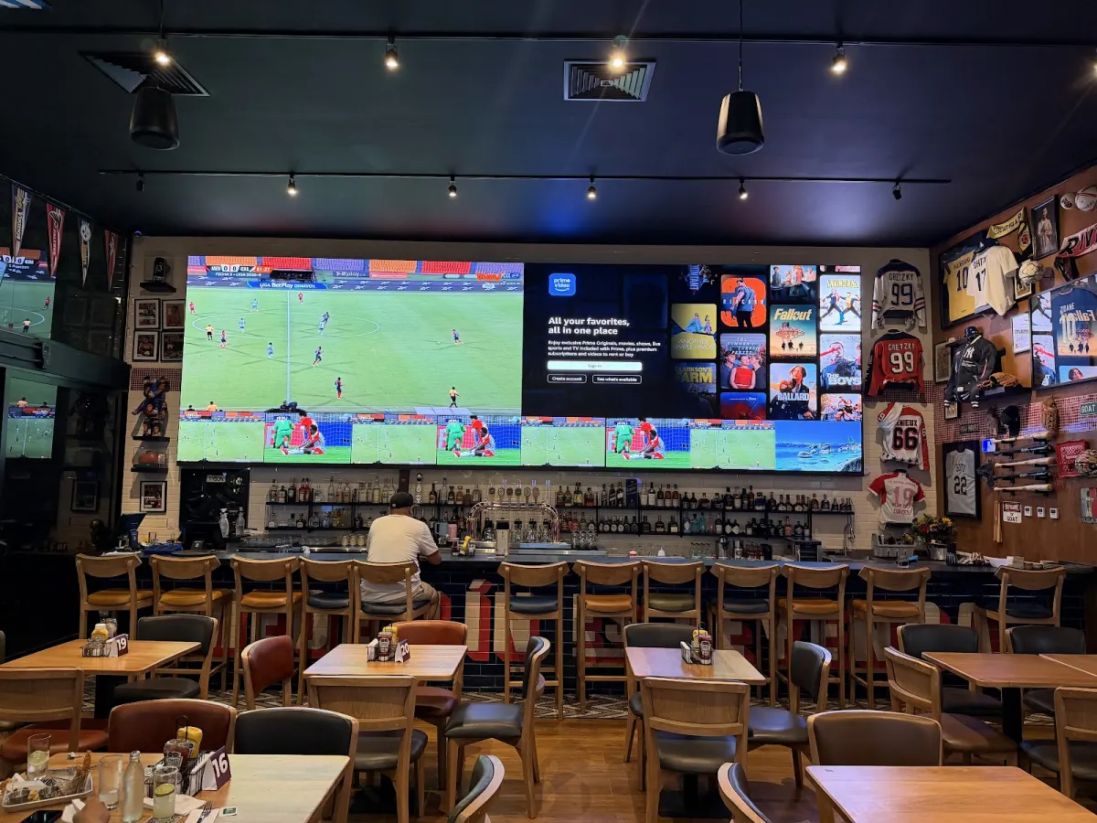 GOAT Sports Bar dining room with a large video wall above the bar, rows of bar stools, wooden tables and hockey and baseball jerseys on the wall