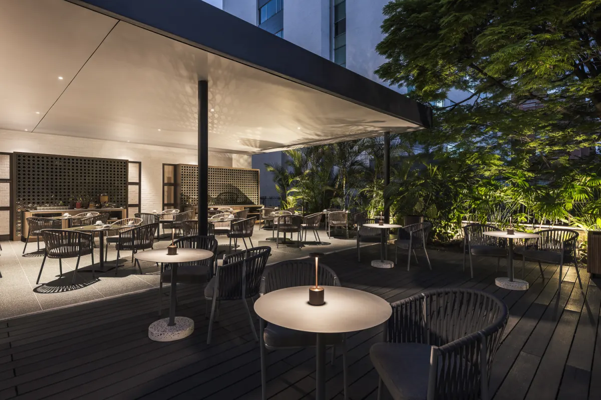 The Hotel Poblado Plaza's outdoor terrace at night, with round tables, woven chairs and table lamps on a timber deck under tropical trees
