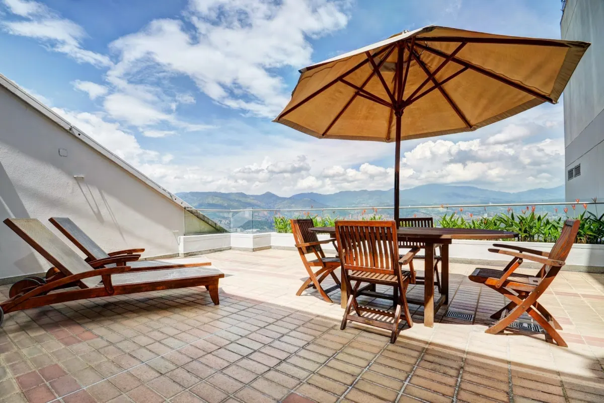 A sunny terrace at Hotel San Fernando Plaza with wooden loungers, a table and chairs under a parasol, and green mountains beyond