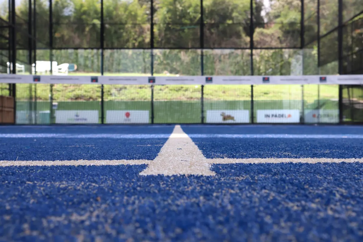 A low view across the blue artificial-grass court at In Pádel Medellín, looking along the white centre line to the net and the glass walls, with In Pádel banners and a lawn beyond