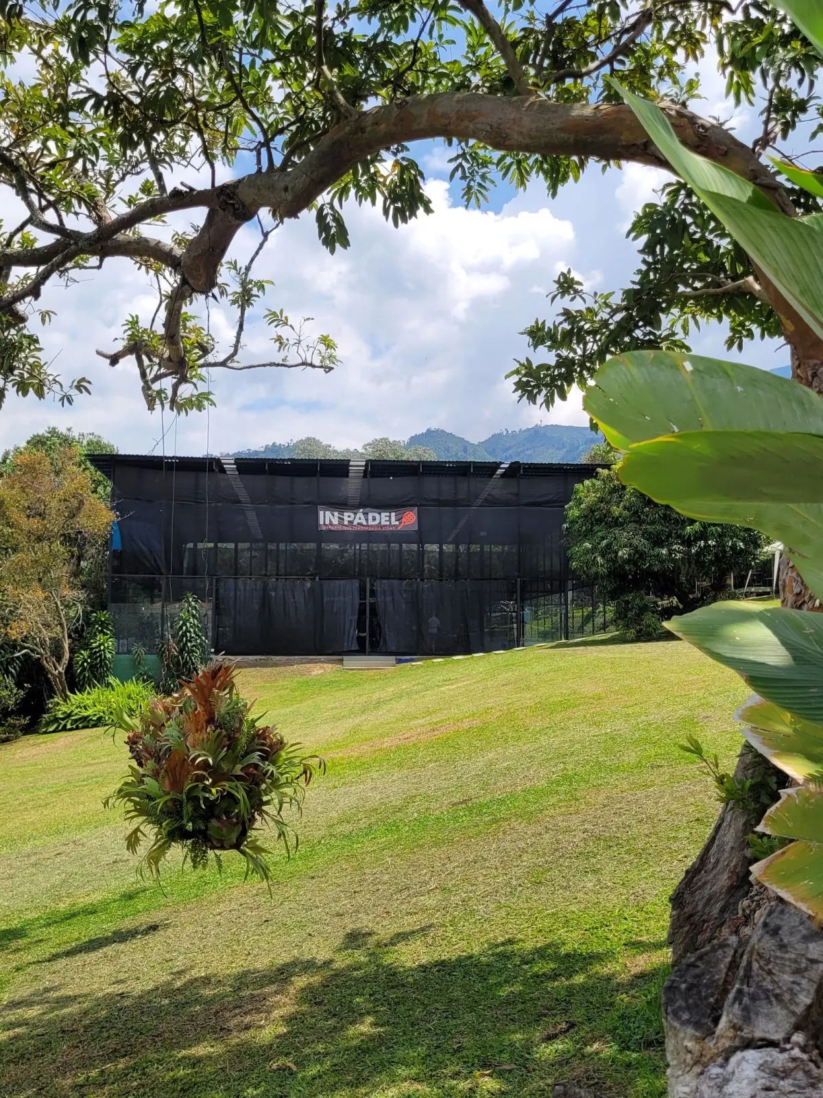 The covered court building at In Pádel Medellín, with an In Pádel banner on its black screening, seen across a sloping lawn under a tree with green hills behind