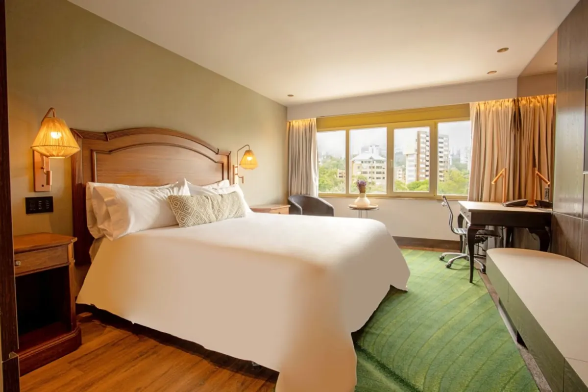 A guest room at the InterContinental Medellin with a double bed, wicker wall lamps, a green rug, a desk and a window over the trees
