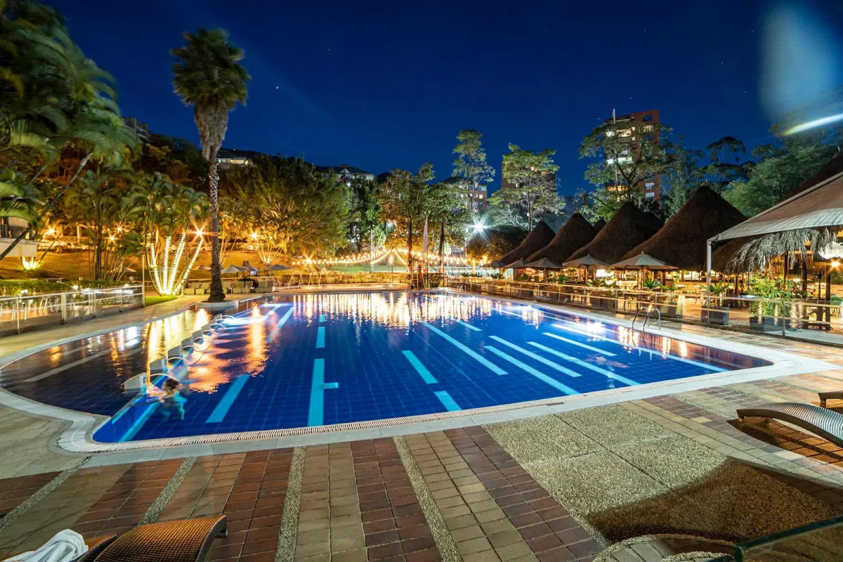The InterContinental Medellin pool at night, lit from below, with palm trees, string lights and thatched cabanas along the far side