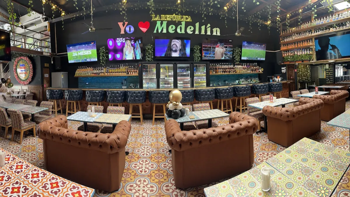 Interior of La República Brunch Sport Bar with a 'Yo ♥ Medellín' neon sign, a row of TVs over the bar, tufted leather sofas and patterned tile floors