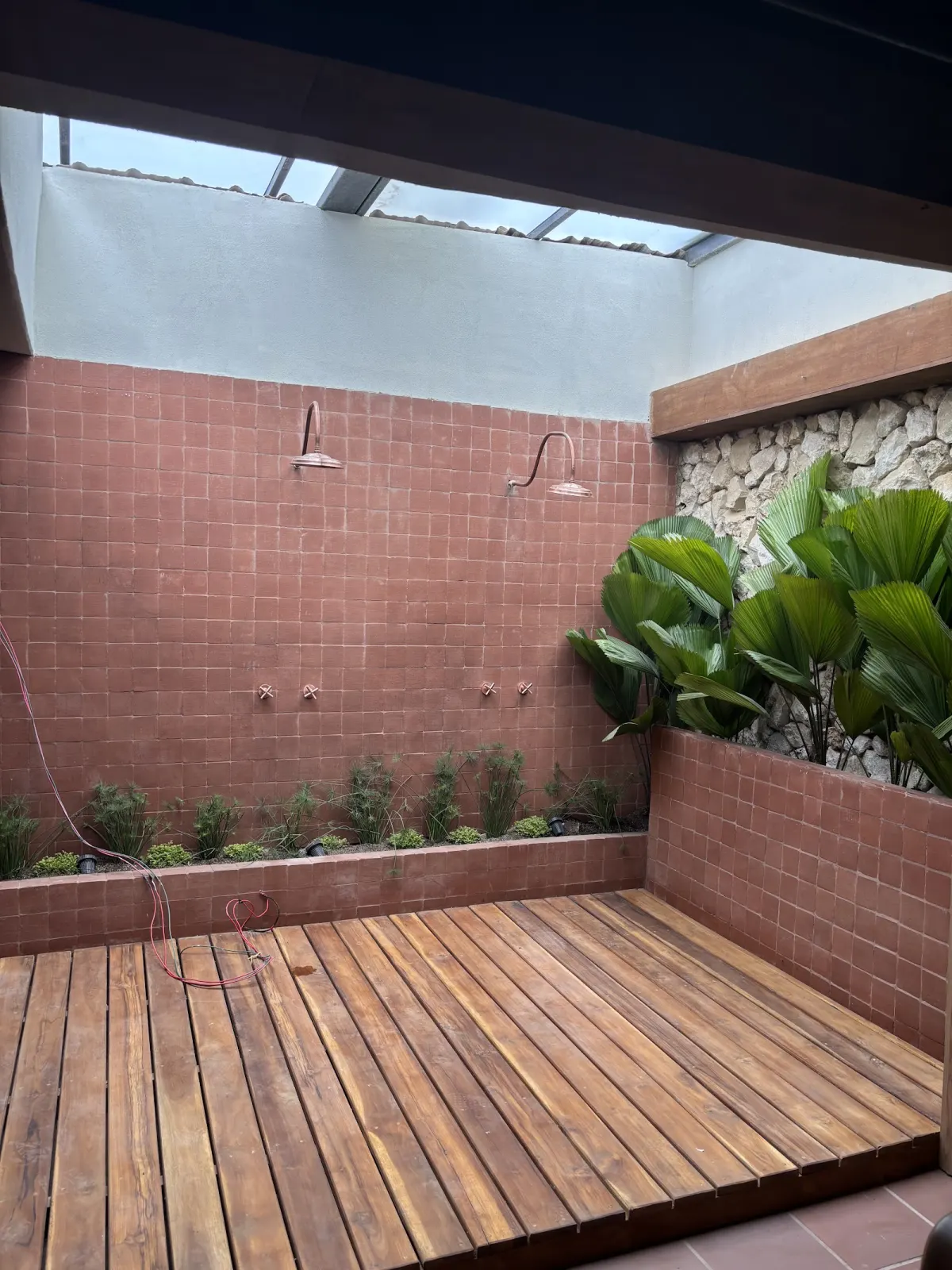 An open-air shower area at MADRE with two copper rain showers on a terracotta-tiled wall, a wooden deck floor and fan palms against a stone wall