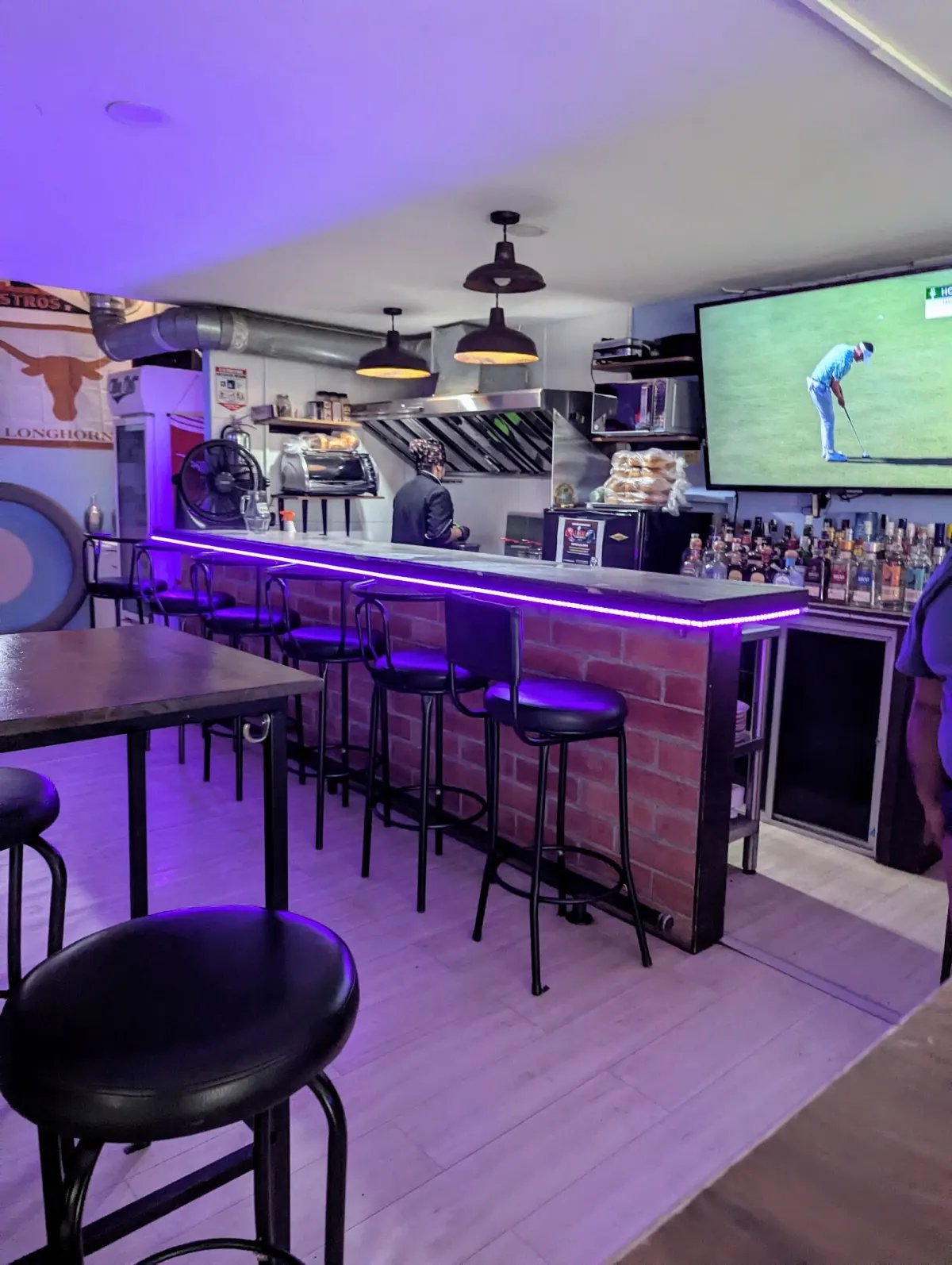 Brick bar counter with purple LED lighting and black stools at Medellín Sports Bar, with a Texas Longhorns flag on the wall and golf on the TV