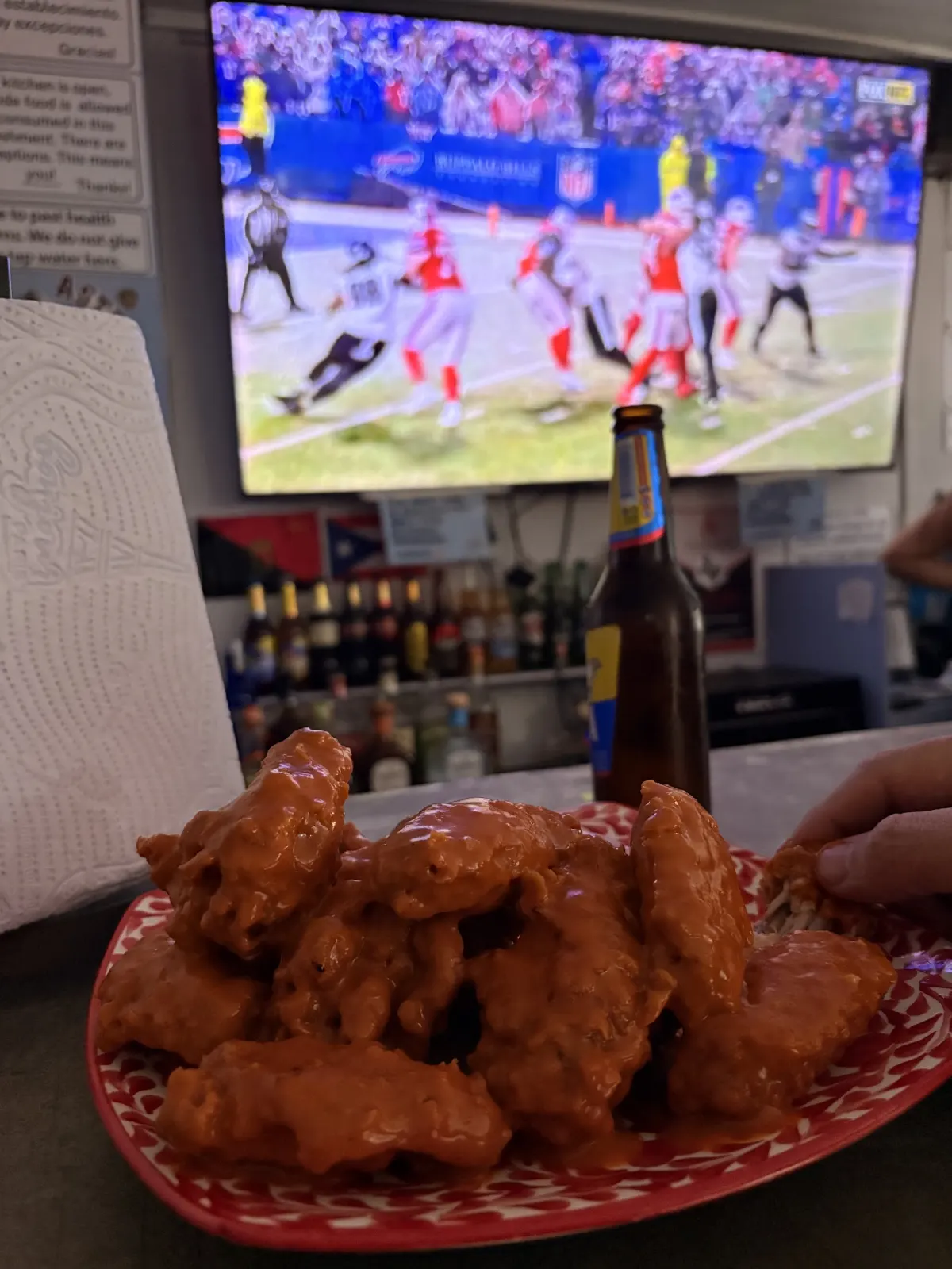 Plate of sauced buffalo wings and a bottle of beer on the bar at Medellín Sports Bar, with an NFL game on the TV behind
