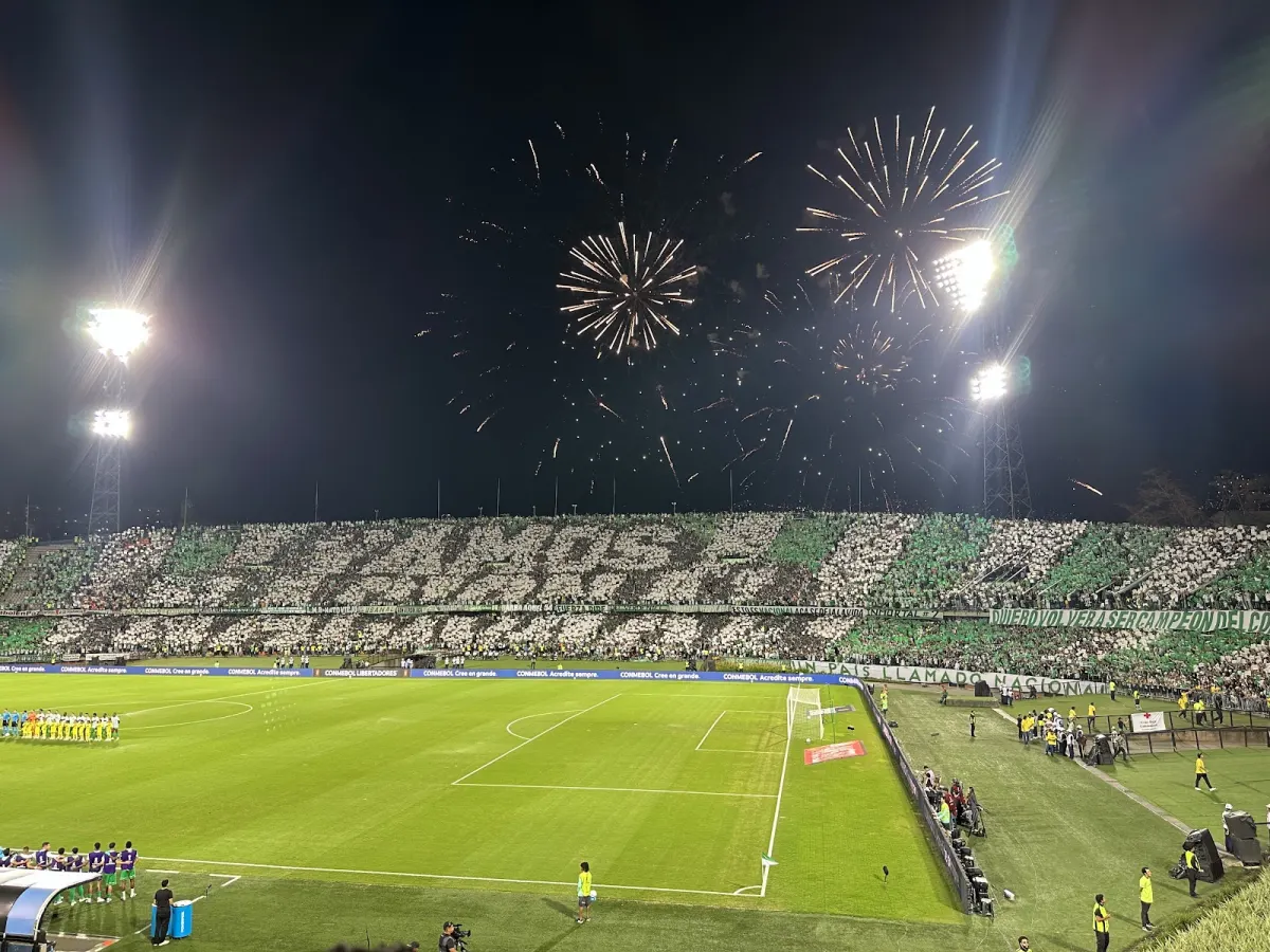 Estadio Atanasio Girardot, where the match-day experience goes: fans in green and white fill the stands of the Estadio Atanasio Girardot at night as fireworks burst over the floodlights and the teams line up on the pitch
