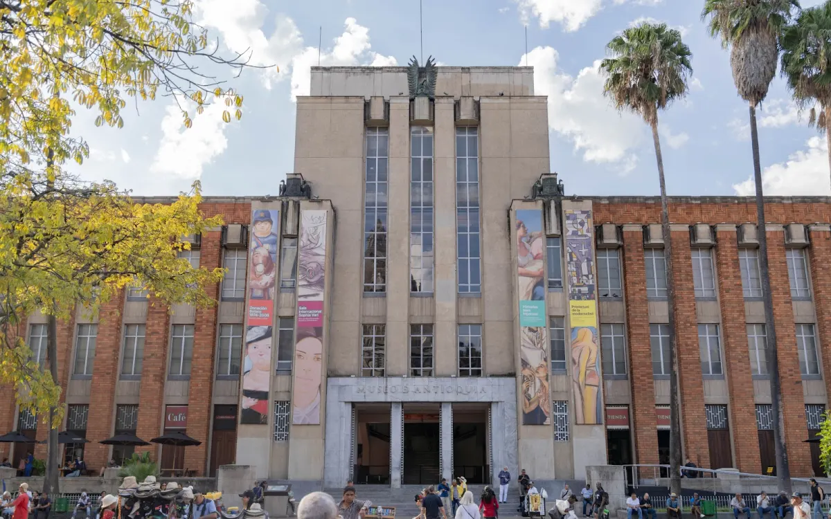 The Museo de Antioquia's stone and brick facade with tall windows and hanging art banners, palm trees and visitors on the steps outside
