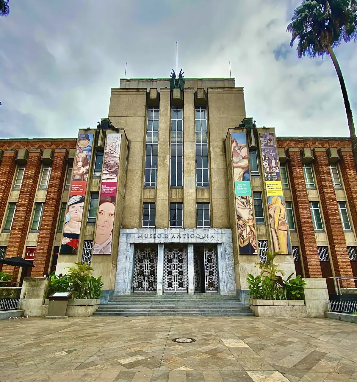 The museum's central entrance with patterned metal doors under the name MUSEO DE ANTIOQUIA, framed by brick wings hung with art banners
