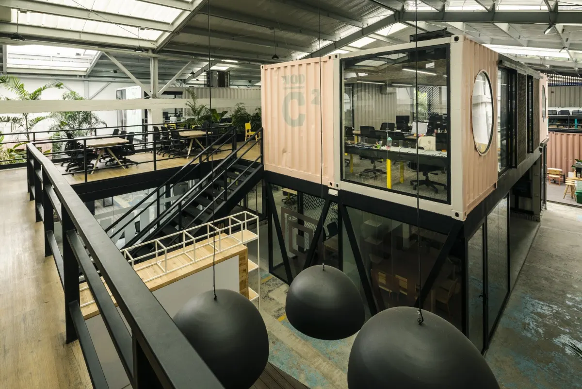 Inside NOI, a glass-fronted shipping-container office with a round window beside a mezzanine of desks and a black steel staircase, under a skylit roof