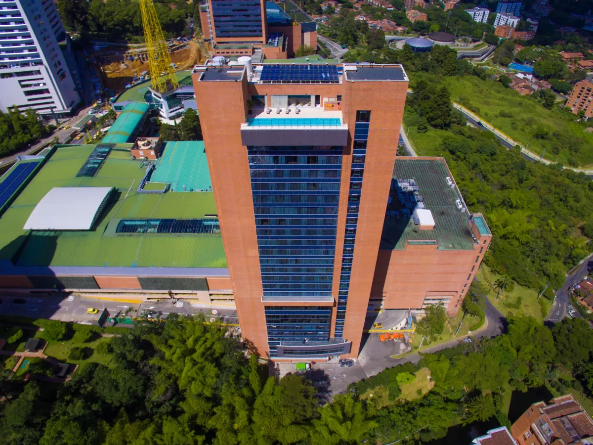 An aerial view of Novotel Medellín El Tesoro, a brick tower with a rooftop pool beside El Tesoro Shopping Park and green hillsides