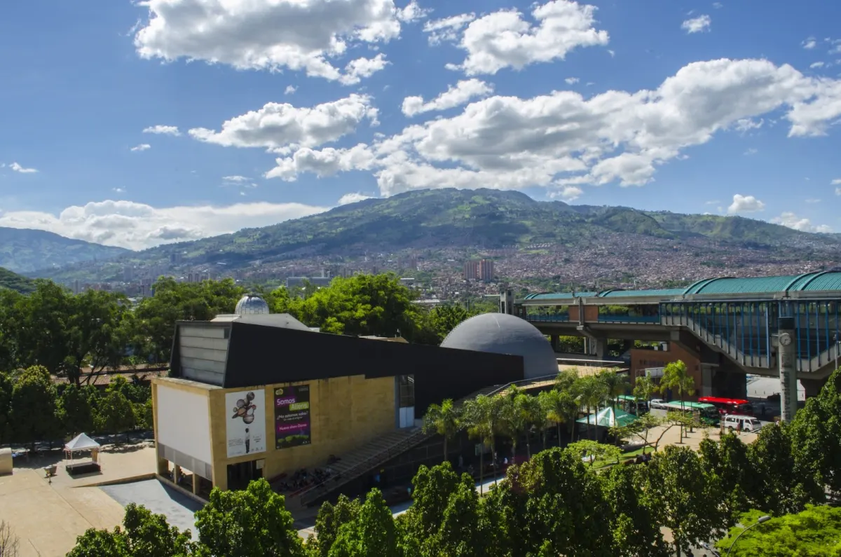 The Planetario de Medellín's building and silver dome in the Parque de los Deseos, seen from above, with green mountains and the city behind under a blue sky