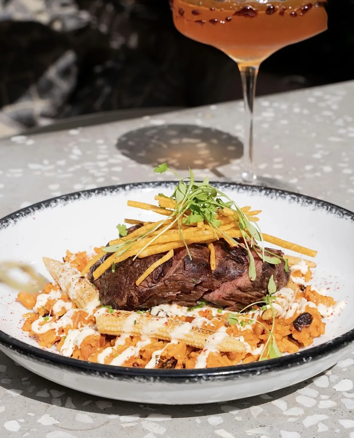 A steak on orange rice with shoestring potatoes and a cocktail behind it, at Posada Salsera