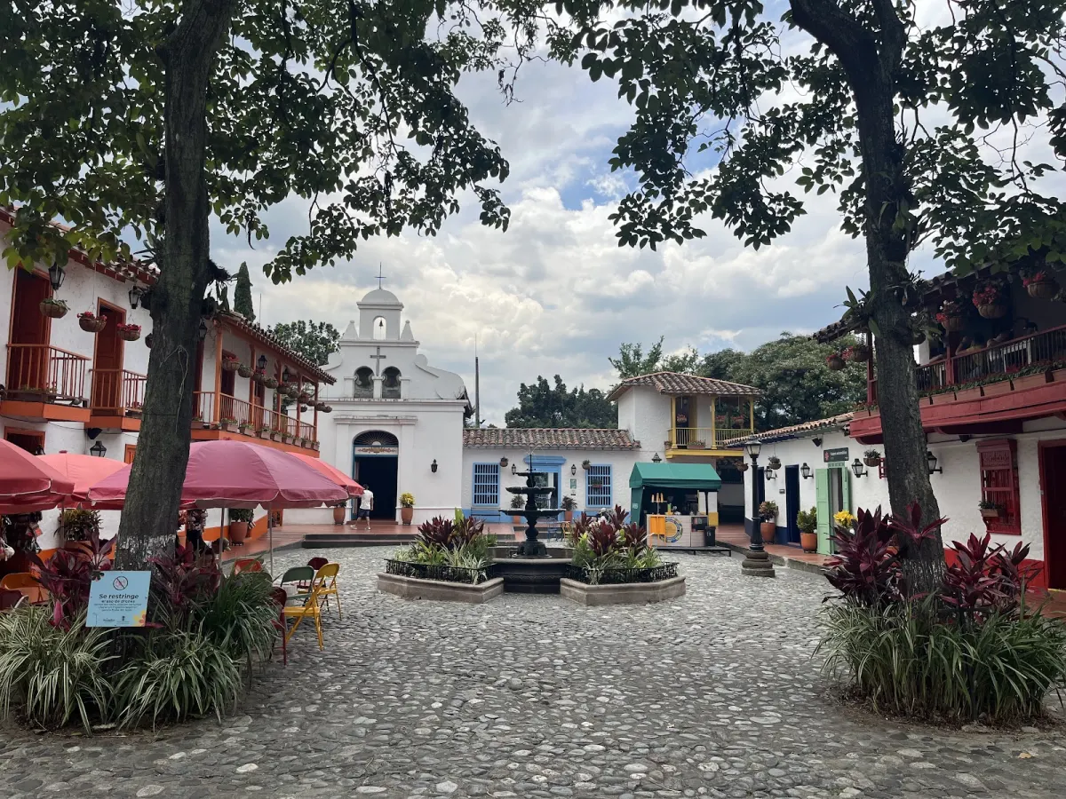 The cobbled main square of Pueblito Paisa, with a fountain, a white church with a bell gable, and whitewashed houses with coloured balconies and flower pots
