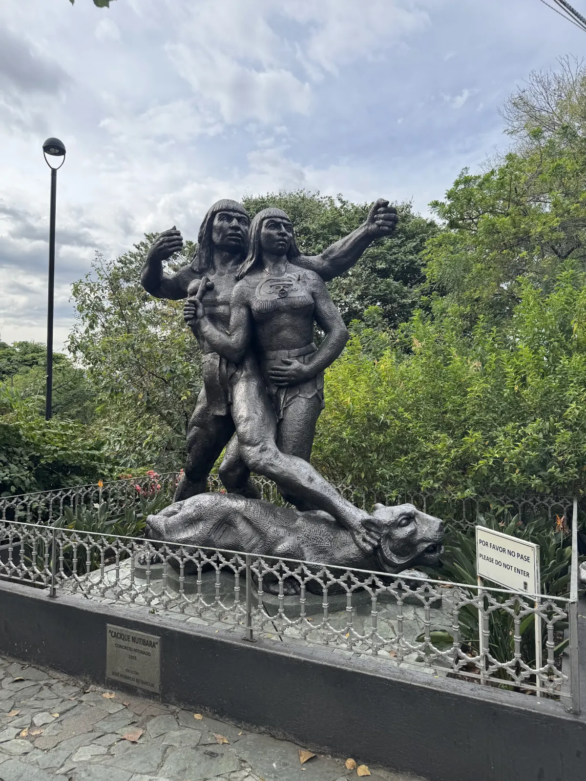 The dark Cacique Nutibara sculpture of two Indigenous figures standing over a puma, behind a low railing with a plaque naming the sculptor José Horacio Betancur
