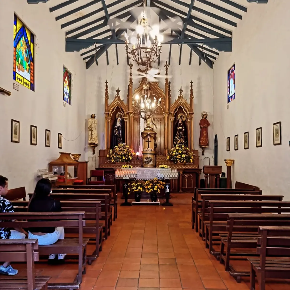 Inside Pueblito Paisa's small white church, with wooden pews, a carved wooden altarpiece, chandeliers and stained-glass windows