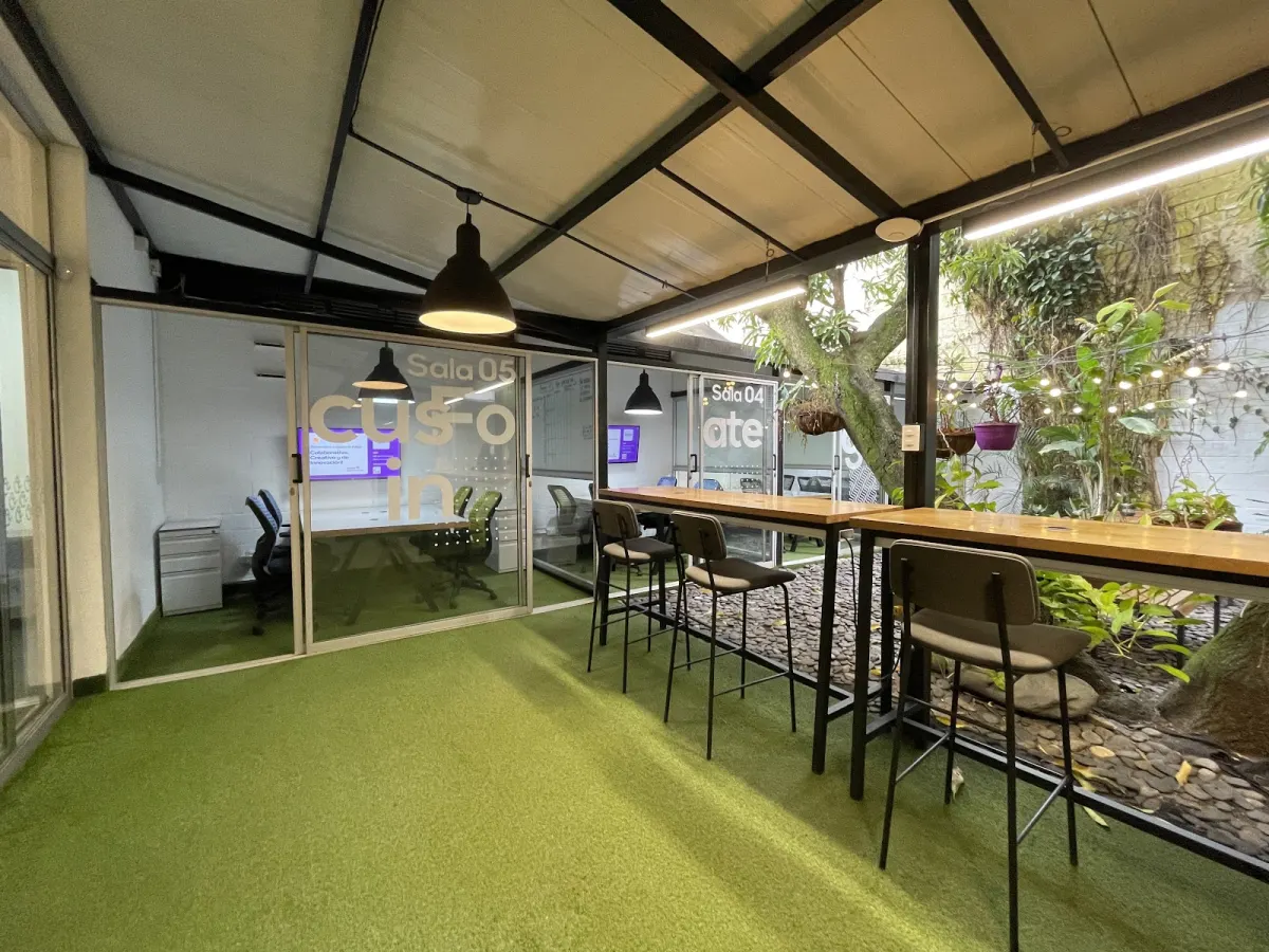 A covered work area at Quokka Astorga with high wooden tables and stools on artificial grass, glass-walled meeting rooms marked Sala 04 and Sala 05, and a tree in the garden alongside