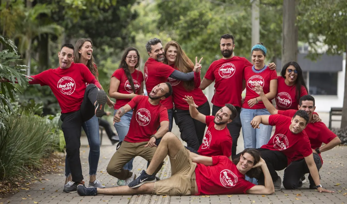 A group of about fourteen people in matching red Real City Tours T-shirts posing playfully on a tree-lined paved path