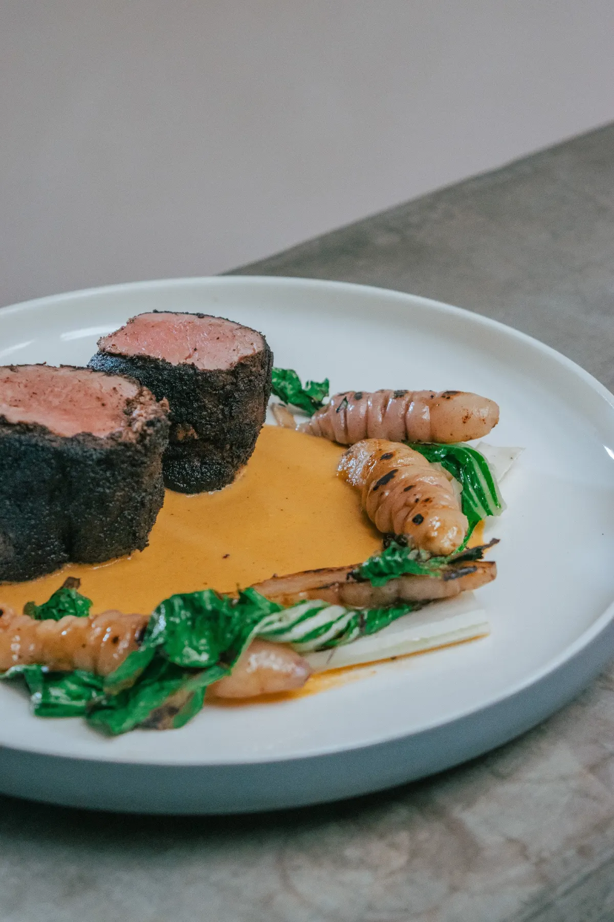 Two slices of dark-crusted beef with an orange sauce, grilled tubers and greens on a white plate at Salón Centro