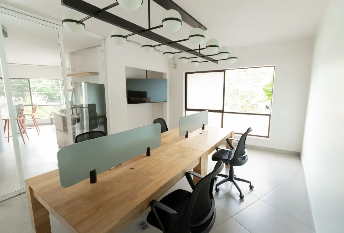 A small room at Semilla with a wooden bench desk split by frosted screens, black office chairs and a wall-mounted screen, with a glass wall onto a kitchen