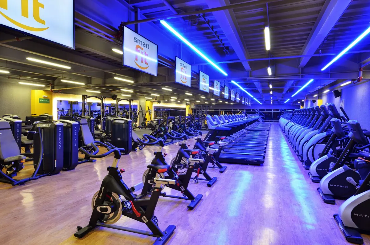 Inside Smart Fit La 10, rows of exercise bikes, treadmills and cross trainers under blue strip lights and Smart Fit screens