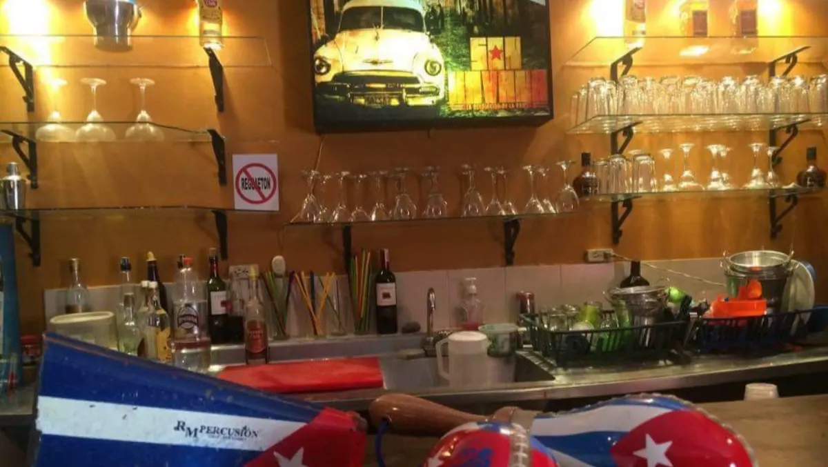 The back bar at Son Havana, with glass shelves of glasses and bottles, a framed picture of a vintage car and a pair of maracas in Cuban-flag colours in the foreground.